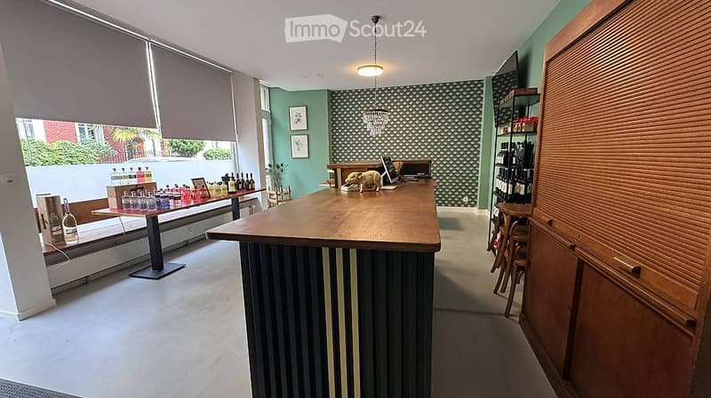 A modern, well-lit bar area with a wooden bar counter, shelves stocked with bottles, a laptop on the counter, and a small table with stools. The space features teal walls, large windows with blinds, and decorative framed pictures.