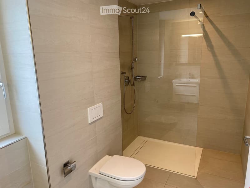 Modern bathroom with a toilet, glass-enclosed shower, and a sink with a faucet. The walls and floor are tiled in a light beige color.