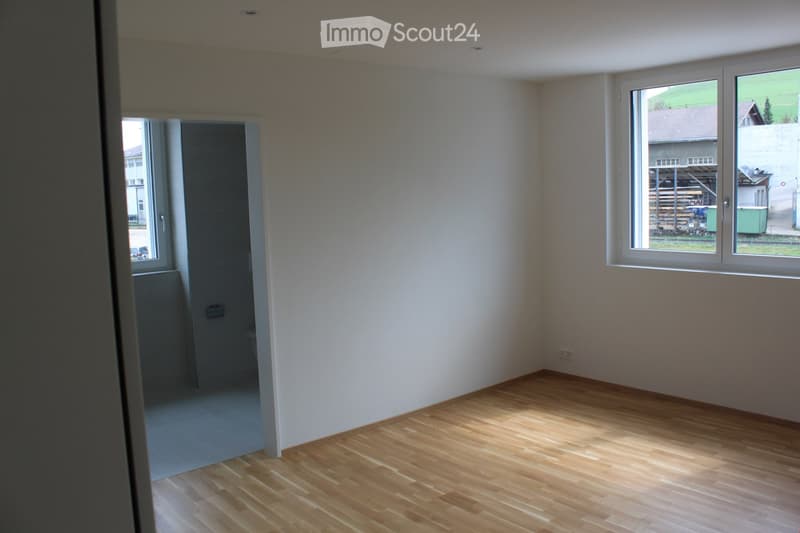 Empty room with light-colored walls and wooden flooring, featuring large windows and a doorway leading to another space.