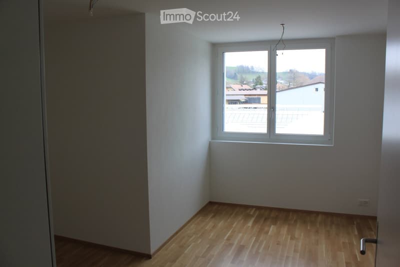Empty room with wooden flooring, white walls, and a large window offering a view of the outside area.