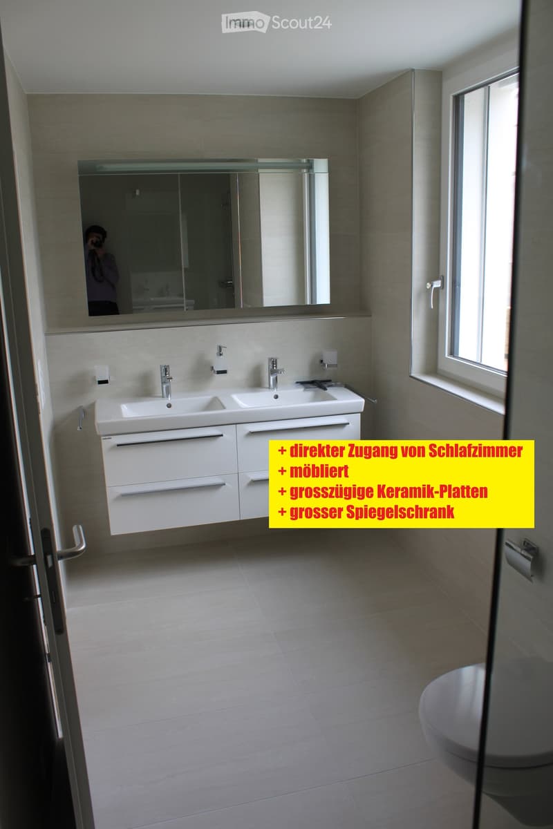 Modern bathroom with double sink, large mirror, ceramic tiles, and a toilet. Features a spacious design with a window and sleek fixtures.