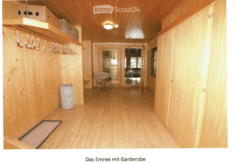 A narrow hallway with wooden walls and flooring, featuring built-in coat racks, a small chair, and a trash bin. The hallway leads to an open doorway with glass panels.