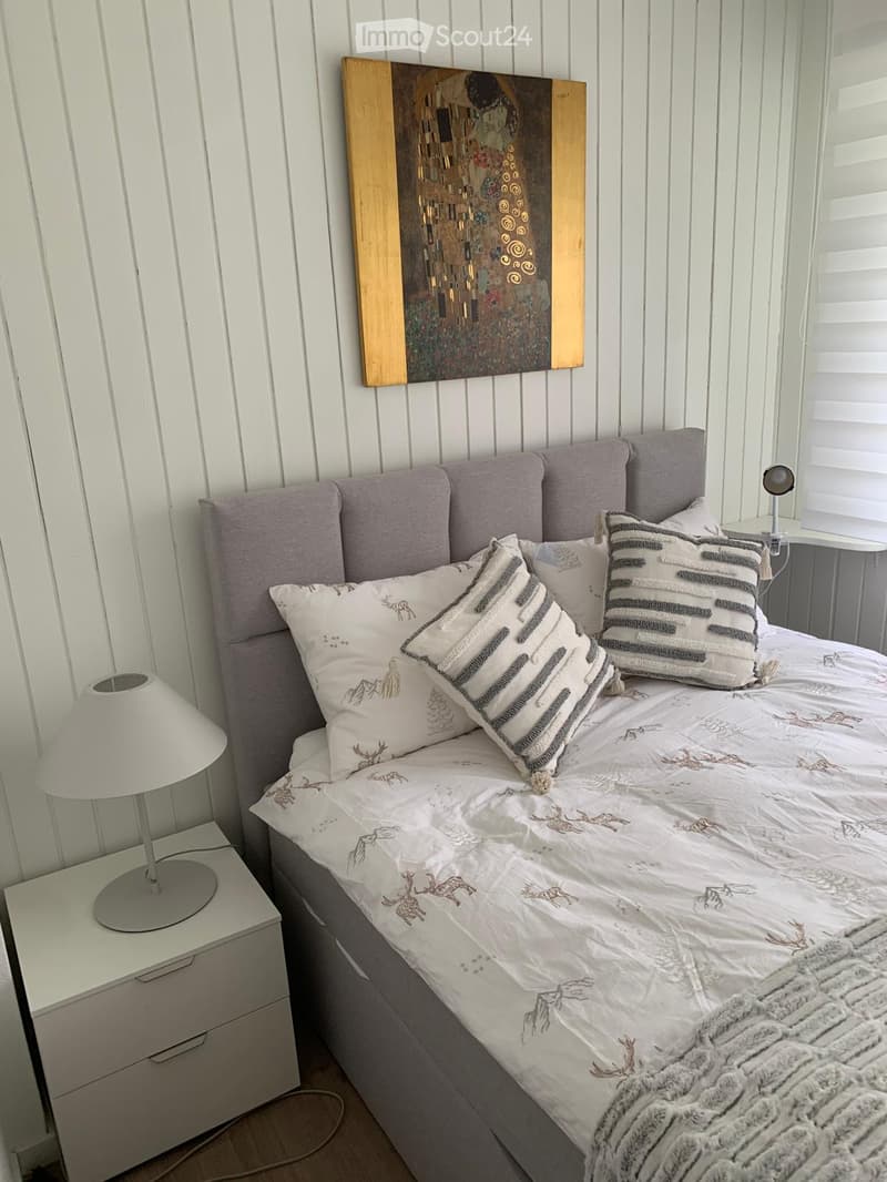 A cozy bedroom with a large bed, grey headboard, decorative pillows, and a soft blanket. A nightstand with a lamp is on the left side, and a painting hangs on the wall above the bed.