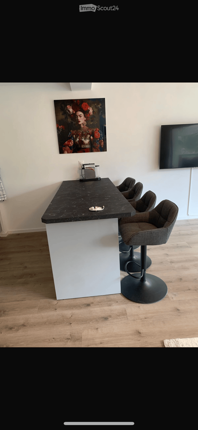 Modern bar area with a black marble countertop, white base, four grey bar stools, a painting on the wall, and a television mounted on the wall.