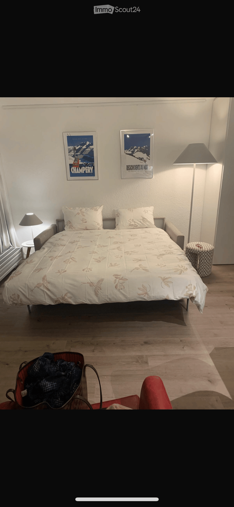 Cozy bedroom with a double bed, nightstand, lamp, and two picture frames on the wall. The room features wooden flooring and a small red chair near the bed.