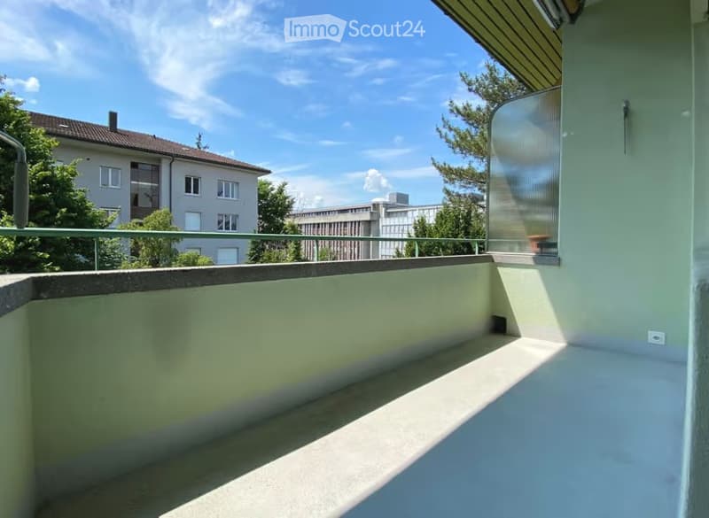 Balcony with a smooth concrete floor, metal railing, and a view of neighboring buildings and greenery.