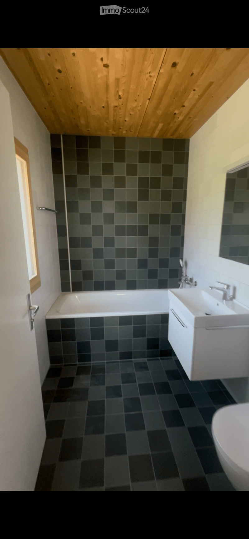 Modern bathroom with dark grey tiles, a white bathtub, a white sink with a chrome faucet, and a wooden slatted ceiling.