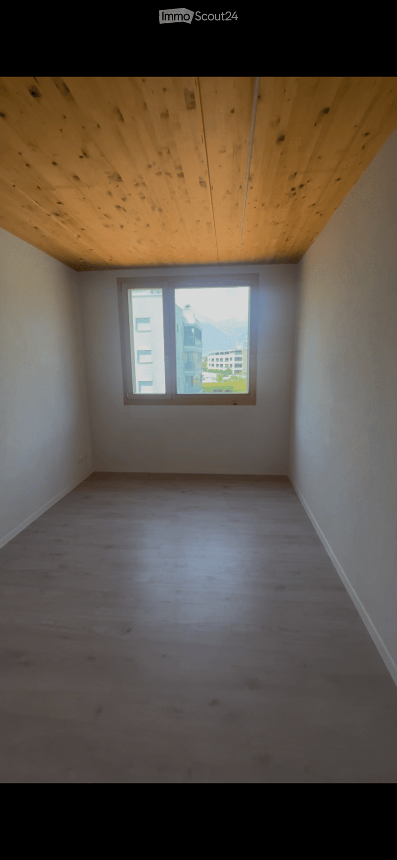 Empty room with wooden floor, white walls, and a single window showing a view of buildings and greenery outside.