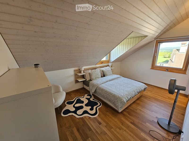 Cozy attic bedroom with wooden slat ceiling, large windows, a bed with patterned bedding, a nightstand, a chair, a fan, and a decorative rug.