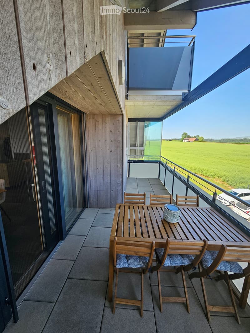 Balcony with wooden table, chairs, and a cup, overlooking a green field.