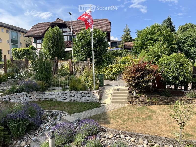 Well-maintained garden with stone walls, lavender bushes, steps leading to a house, and a flagpole with a red and white flag.