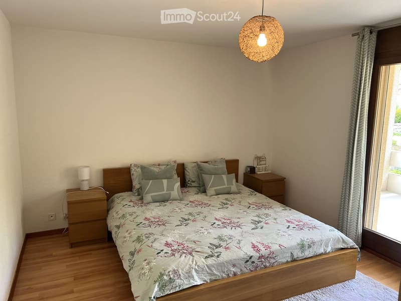 Cozy bedroom with wooden flooring, a neatly made bed with floral bedding, two nightstands with lamps, and a large window with curtains leading to a balcony.