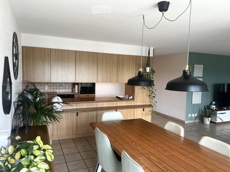 A modern kitchen and dining area with light wooden cabinets, a wooden dining table with white chairs, a fan, and a TV screen in the background.