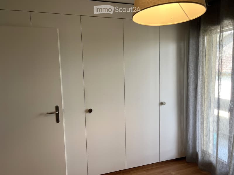 Empty room with light-colored walls, wooden floor, white sliding doors, and a window with sheer curtains.