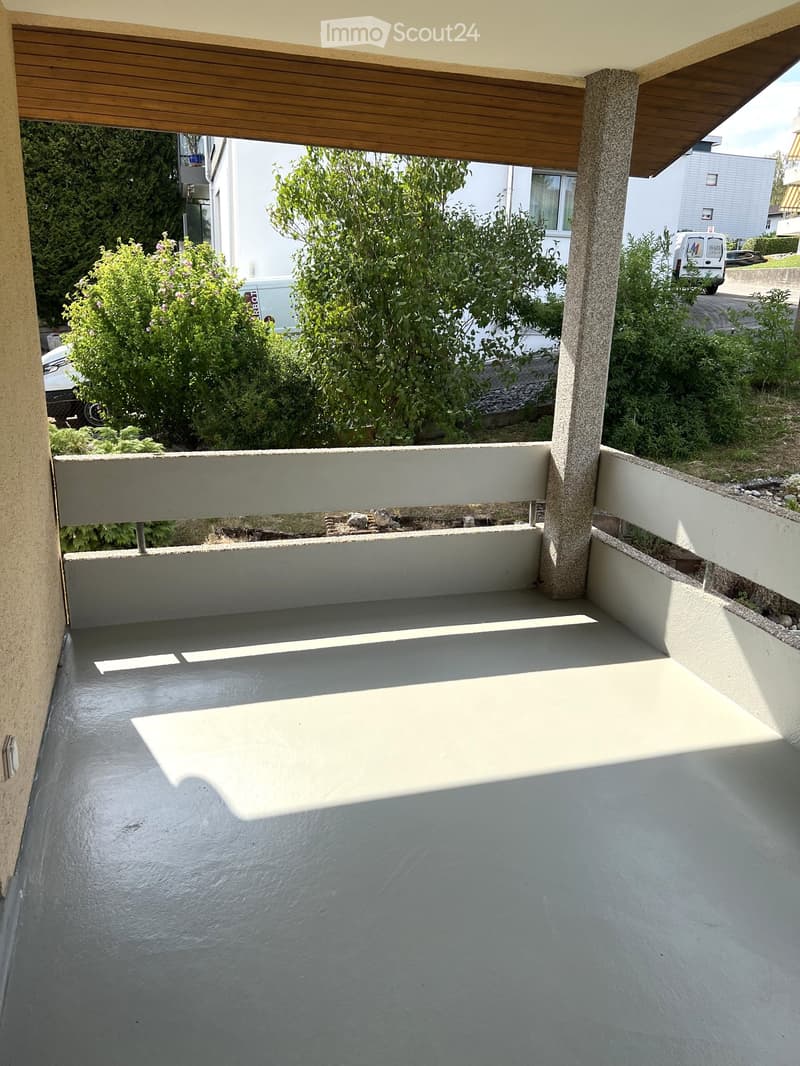 Empty balcony with grey concrete floor, white railing, and wooden ceiling supported by a concrete pillar. The view shows greenery and neighboring buildings.