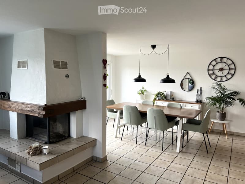 Modern living and dining area with a fireplace, dining table, chairs, and decorative items including a large wall clock and a potted plant.