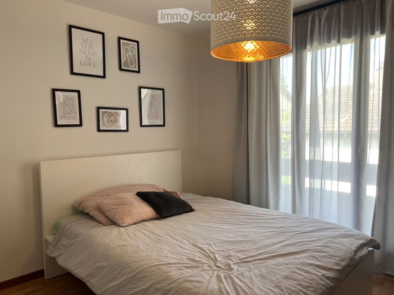 A modern bedroom with a large bed, headboard, pillows, and a hanging lamp. The walls are decorated with several picture frames, and sheer curtains cover the windows.