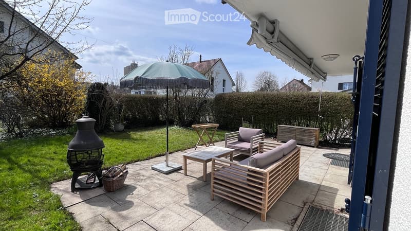 Outdoor patio with wooden furniture, including a sofa, chairs, and a coffee table, along with a large umbrella, a fire pit, and a small table. The area is surrounded by a well-maintained lawn and hedges.