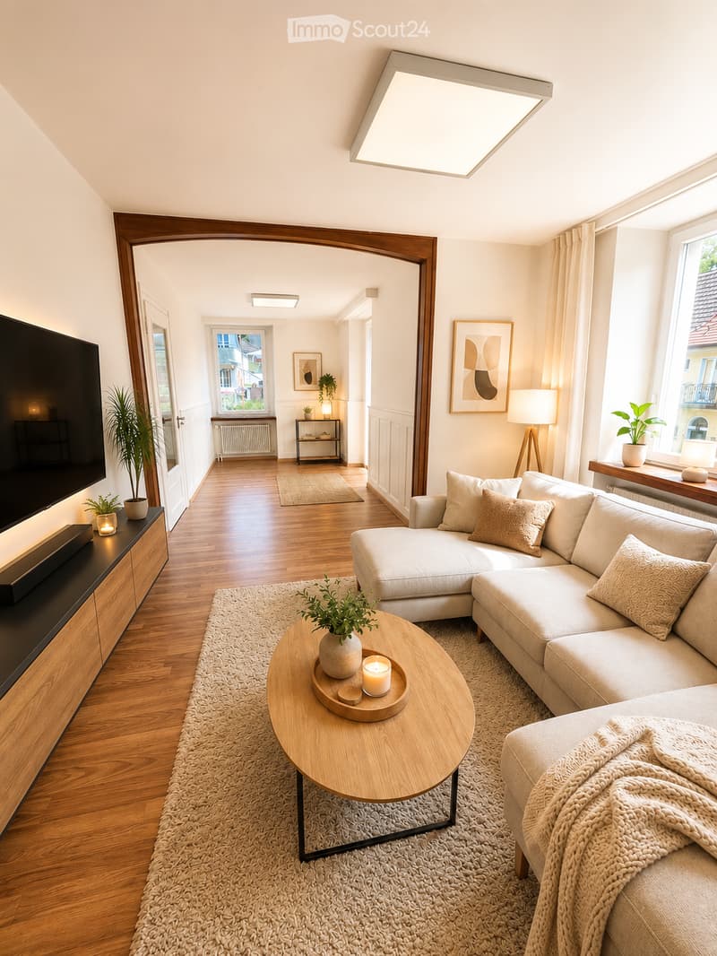 Modern living room with a light-colored sectional sofa, wooden coffee table, floor lamp, side table, and television. The room features wooden flooring, large windows, and decorative plants.