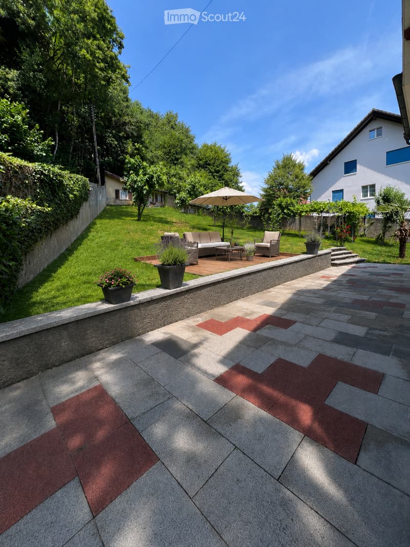 Well-maintained garden with a raised patio, outdoor seating area, umbrella, potted plants, and a stone pathway.