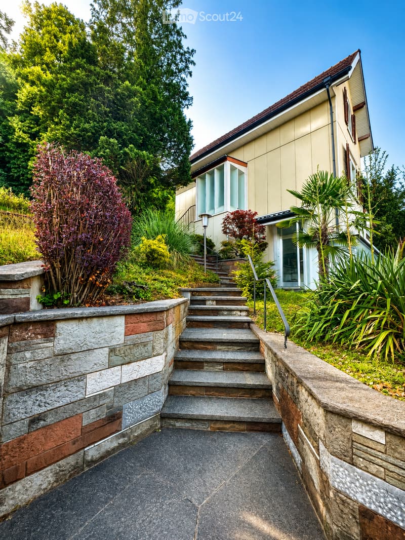 A stone staircase leads up to a modern house with large windows and a well-maintained garden featuring various plants and shrubs.
