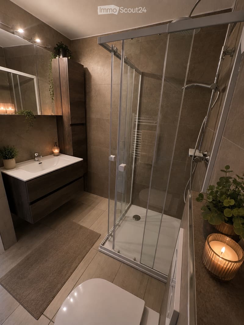 Modern bathroom with a glass-enclosed shower, a white bathtub, a sink with a mirror, and decorative candles and plants.