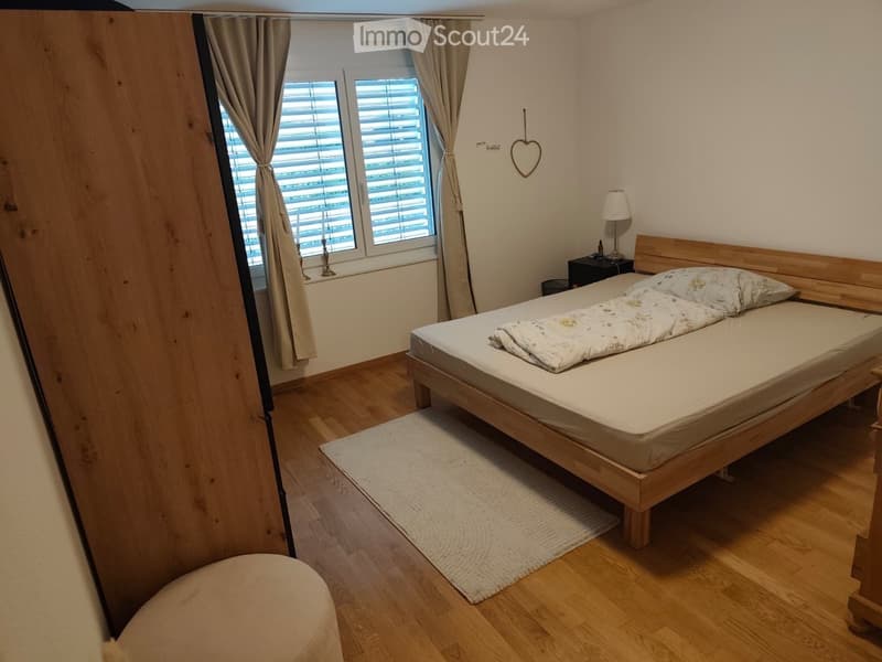Cozy bedroom with wooden flooring, a bed with a patterned duvet, a nightstand with a lamp, and a window with beige curtains.