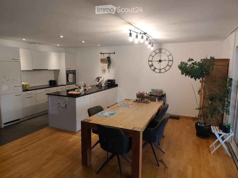 Modern kitchen with white cabinets, black countertops, a central island, a dining table with chairs, and a large wall clock. The floor is wooden, and there are plants and a small stool near the window.