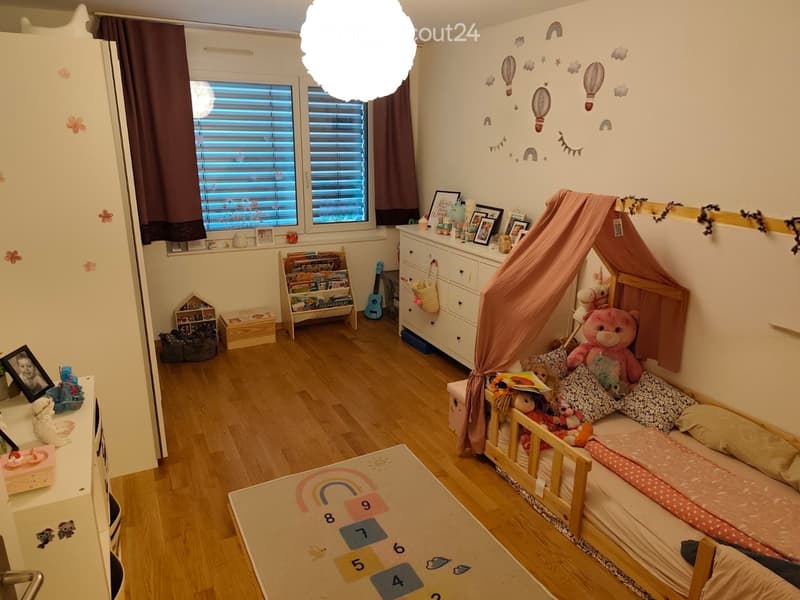 A well-lit children's bedroom with a wooden bed, colorful pillows, a teddy bear, a bookshelf, a guitar, and a toy house. The room features a large window with blinds, a rug with numbers, and various decorations including wall decals and framed photos.