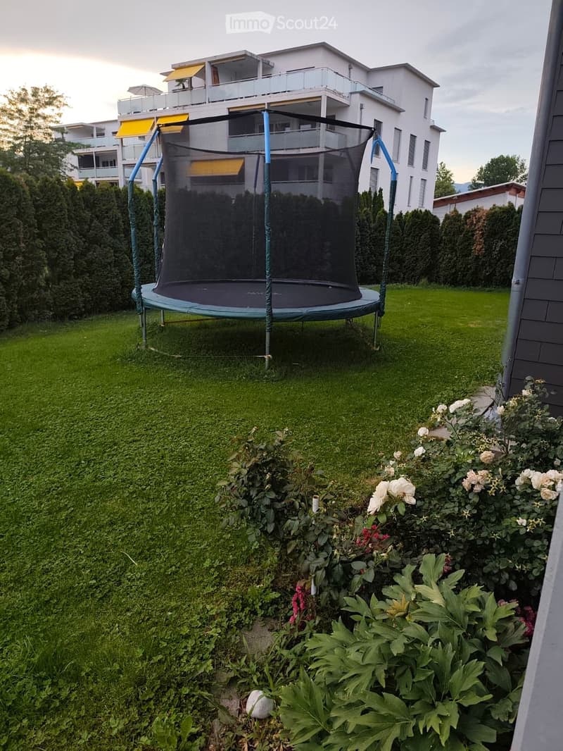 A well-maintained garden with a trampoline, surrounded by green grass, bushes, and flowering plants. A multi-story residential building with balconies is visible in the background.