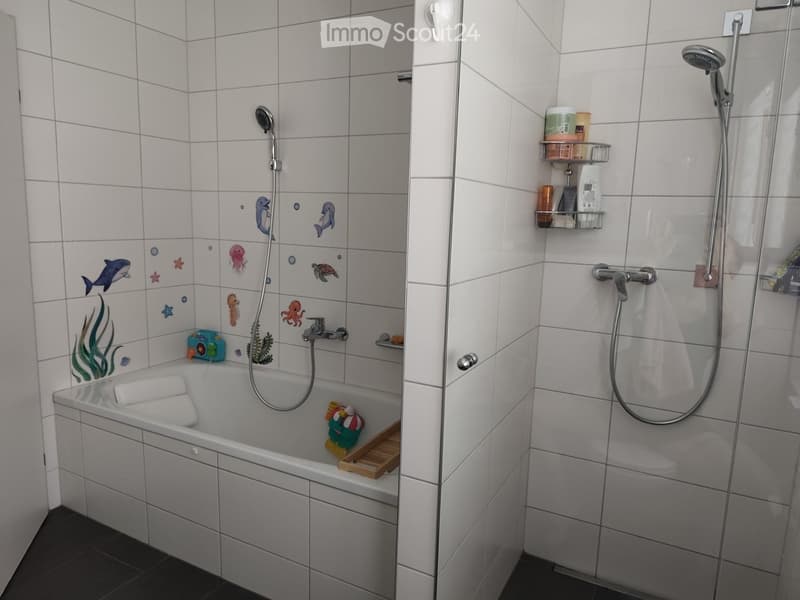 Modern bathroom with a bathtub and separate shower area. The bathtub features a decorative marine-themed tile design, and the shower area includes a glass door, a towel, and a shelf with toiletries.