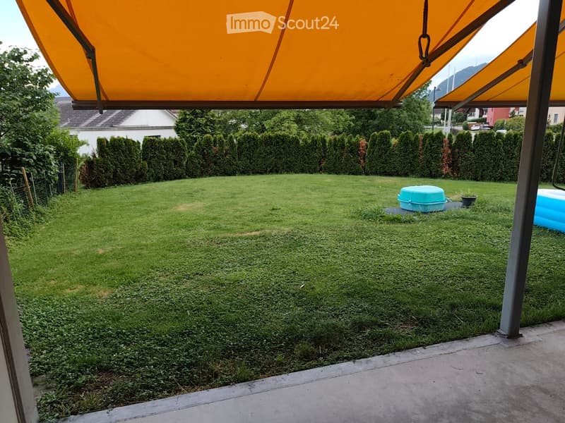 Well-maintained lawn with a neatly trimmed hedge border, a small blue container, and a covered patio area with orange awnings.
