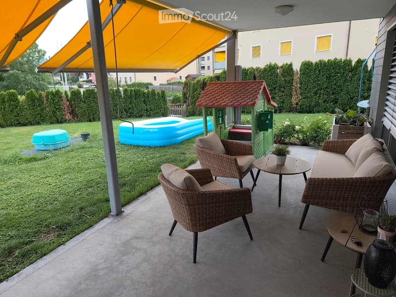 Balcony with wicker furniture, including a sofa, chairs, and small tables, with a view of a backyard featuring a playhouse, inflatable pool, and greenery.