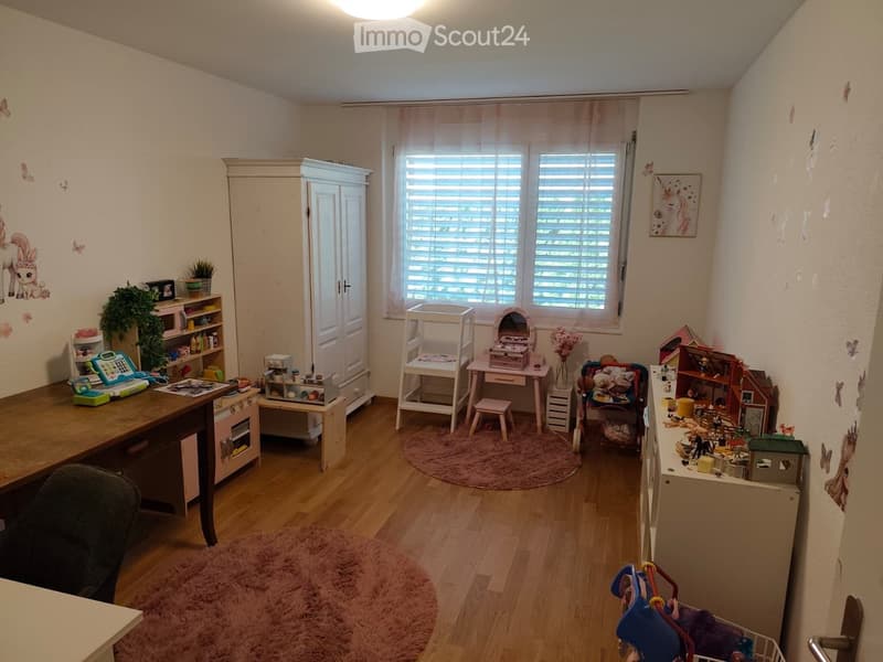 A well-lit children's room with wooden flooring, a large window with blinds, and various toys and furniture including a desk, chair, cabinet, stool, small table, mirror, and multiple toy sets.