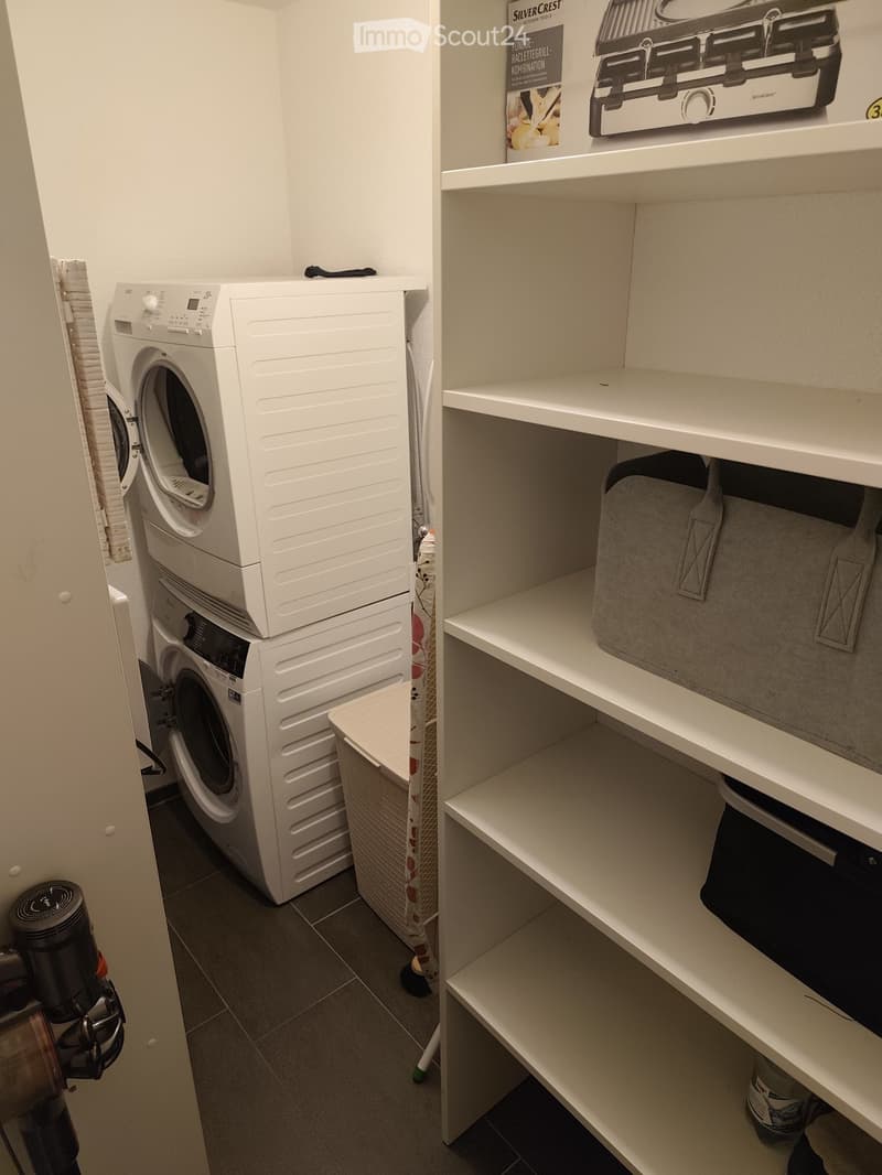 A compact bathroom with a stacked washing machine and dryer unit, white shelves, and a book on top.