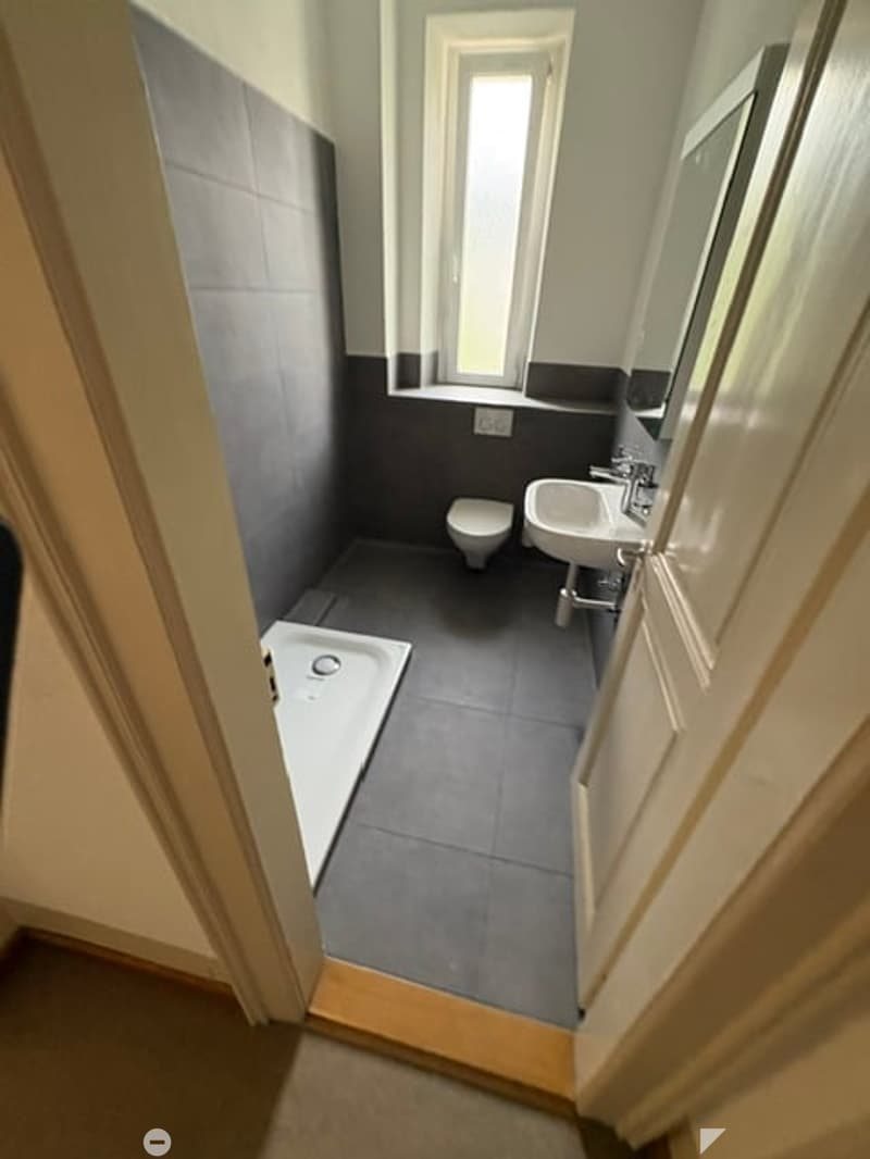 Modern bathroom with grey tiles, a white sink, a toilet, a bathtub, and a mirror above the sink. The room has a window providing natural light.