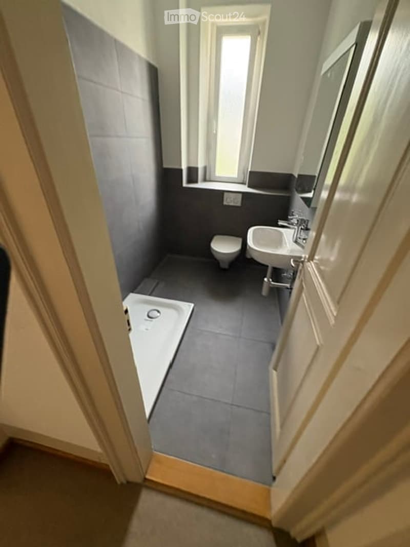 Modern bathroom with grey tiles, a white sink, a toilet, a bathtub, and a mirror above the sink. The room has a window providing natural light.
