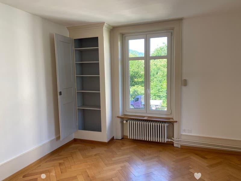 Empty room with wooden herringbone flooring, white walls, a window with a radiator below, and an open built-in cabinet with empty shelves.