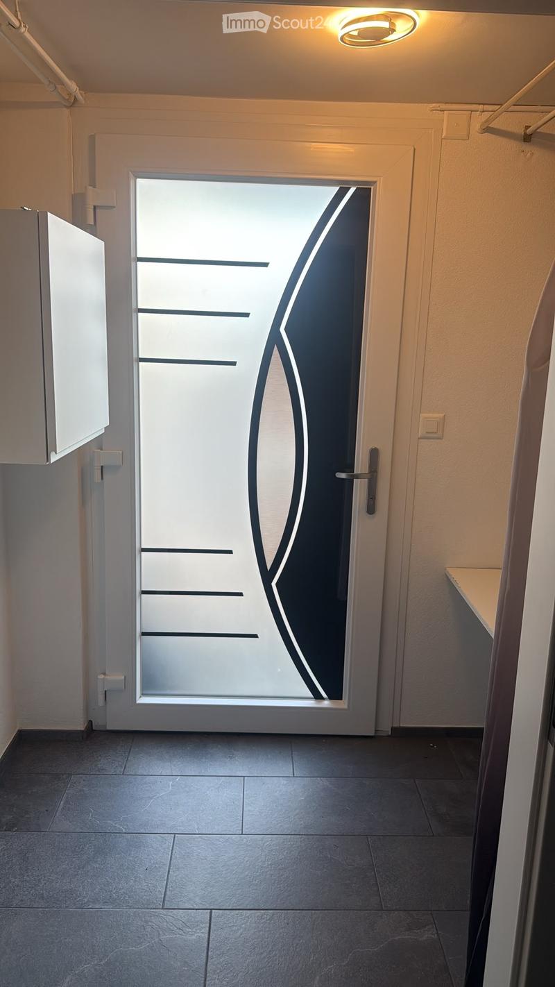 Modern hallway with dark tiled floor, white walls, and a large glass door with a black geometric design. A round ceiling light is visible above the door.