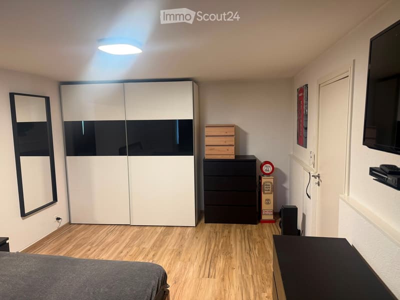 Modern bedroom with a grey bed, white wardrobe, black dresser, mirror, small table, and mounted TV.