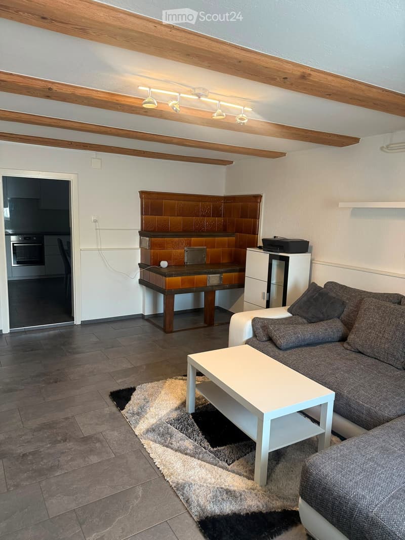 Living room with a grey sofa, white coffee table, wooden table, and an open kitchen area with an oven and cabinets.
