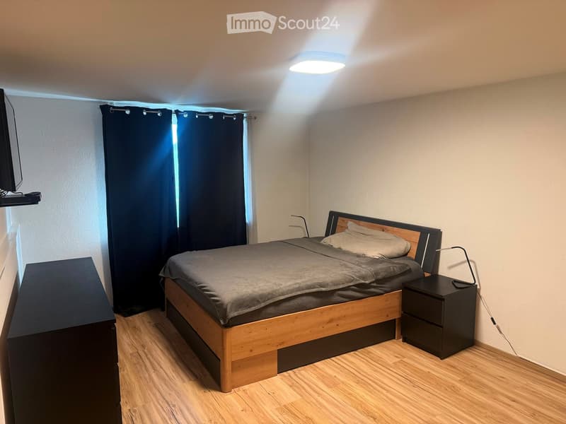 A modern bedroom with a wooden bed, nightstand, lamp, and wall-mounted TV. The room features wooden flooring and dark curtains on the window.