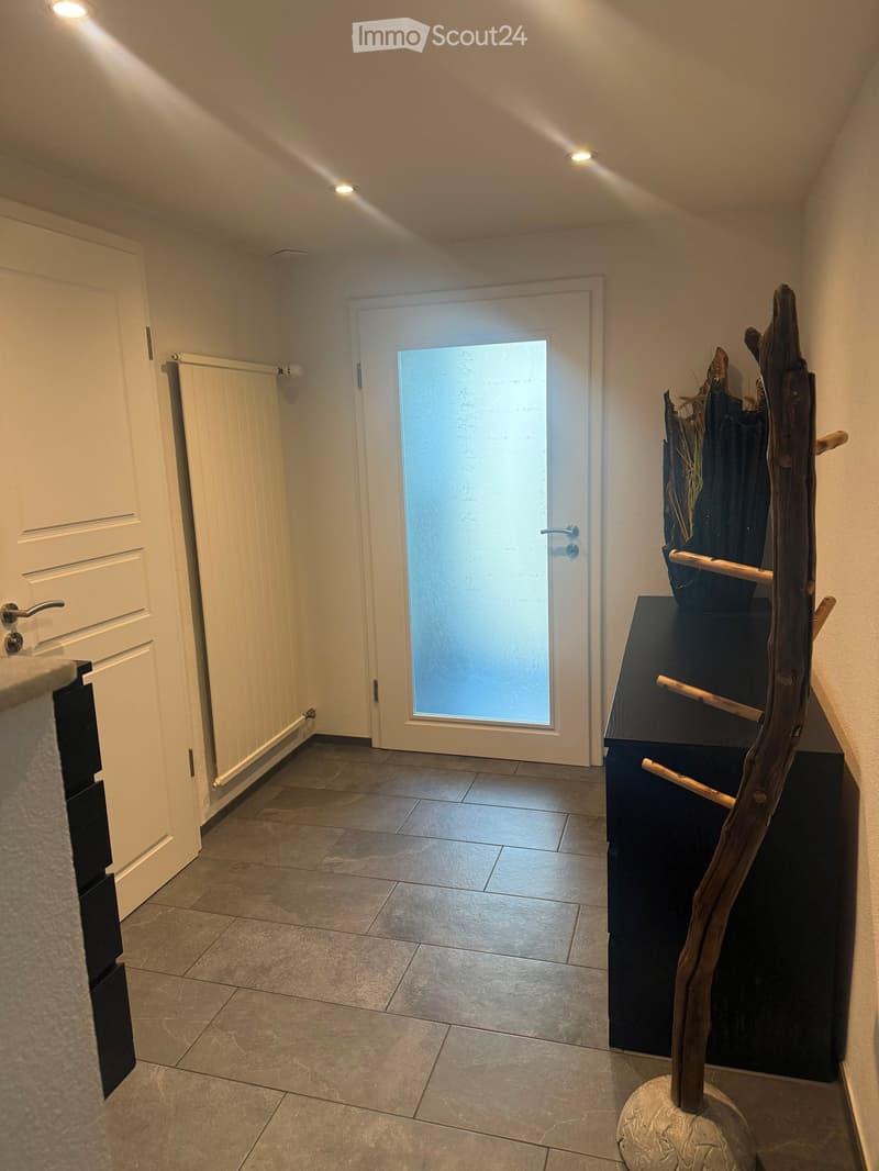 A narrow hallway with tiled flooring, white walls, and recessed lighting. Features a white door on the left and a frosted glass door at the end leading outside. A small black table with a decorative wooden sculpture is placed against the right wall.