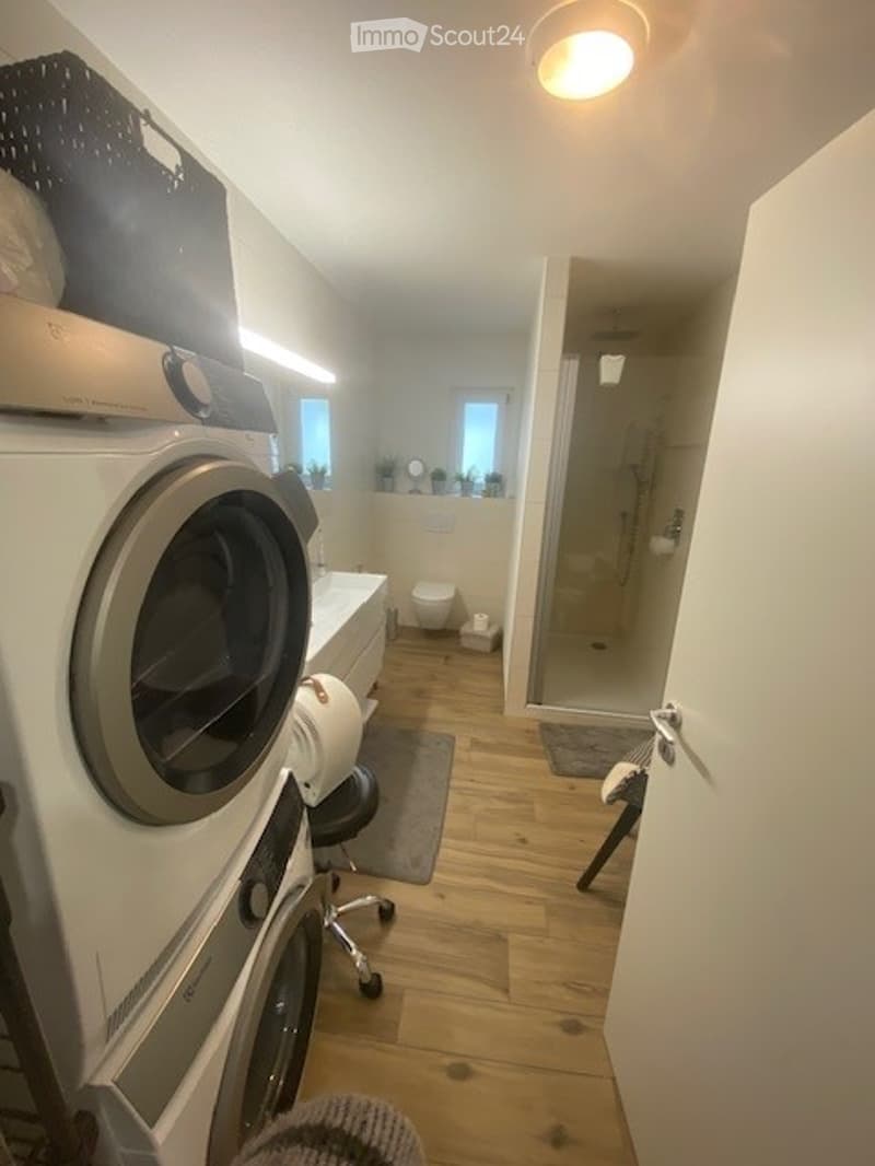 Modern bathroom with a washing machine, toilet, sink, and a glass-enclosed shower. Wooden flooring and white walls.