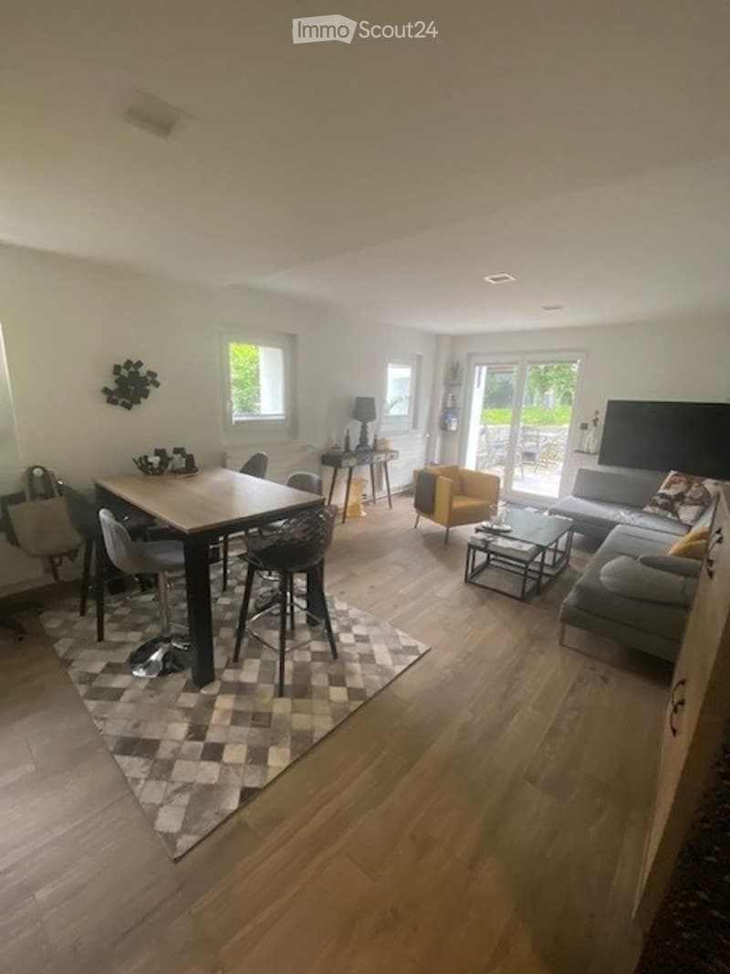 Bright, open-concept living and dining area with hardwood flooring, large windows, and a sliding glass door leading to a balcony. Features a grey sectional sofa, yellow armchair, coffee table, dining table with four chairs, side table with lamp, and wall