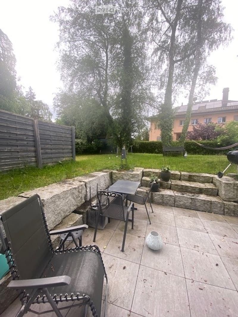 Outdoor patio area with stone steps, a small table, chairs, and a decorative pot. The space is surrounded by a wooden fence and trees, with a grassy lawn and a view of neighboring houses.