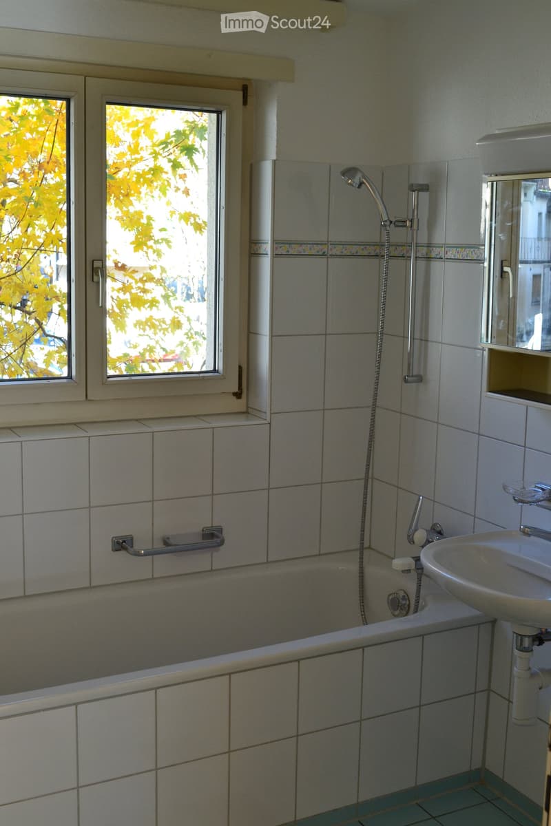 Bathroom with a bathtub, sink, mirror, and handrail. The room has tiled walls and a window with yellow foliage visible outside.