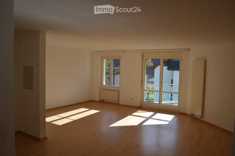 Empty room with wooden flooring, large windows, and a sliding glass door leading to a balcony. Light-colored walls and a radiator are visible.
