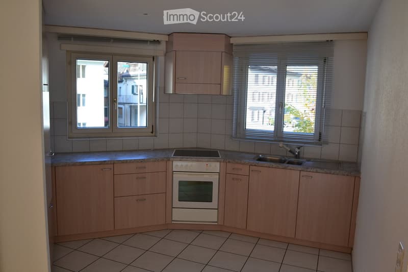Modern kitchen with light wooden cabinets, granite countertop, sink, and built-in oven. Windows with blinds provide natural light.