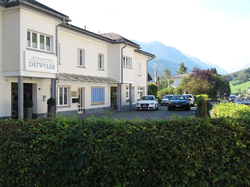 A two-story white building with multiple windows and a sign reading "INTERCOIFFURE DÄTWYLER" above the entrance. Several cars are parked in front, and there is a well-maintained hedge in the foreground. The background features a scenic mountain view.